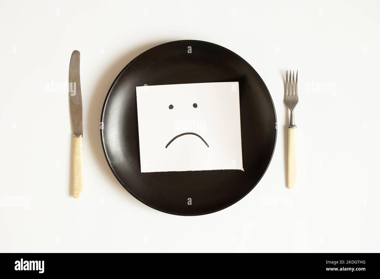 Ein trauriges Emoticon wird auf Papier gezeichnet, liegt auf einem Teller auf weißem Hintergrund, Traurigkeit Traurigkeit und kein Essen Stockfoto