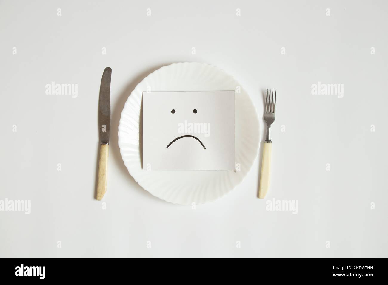 Ein trauriges Emoticon wird auf Papier gezeichnet, liegt auf einem Teller auf weißem Hintergrund, Traurigkeit Traurigkeit und kein Essen Stockfoto
