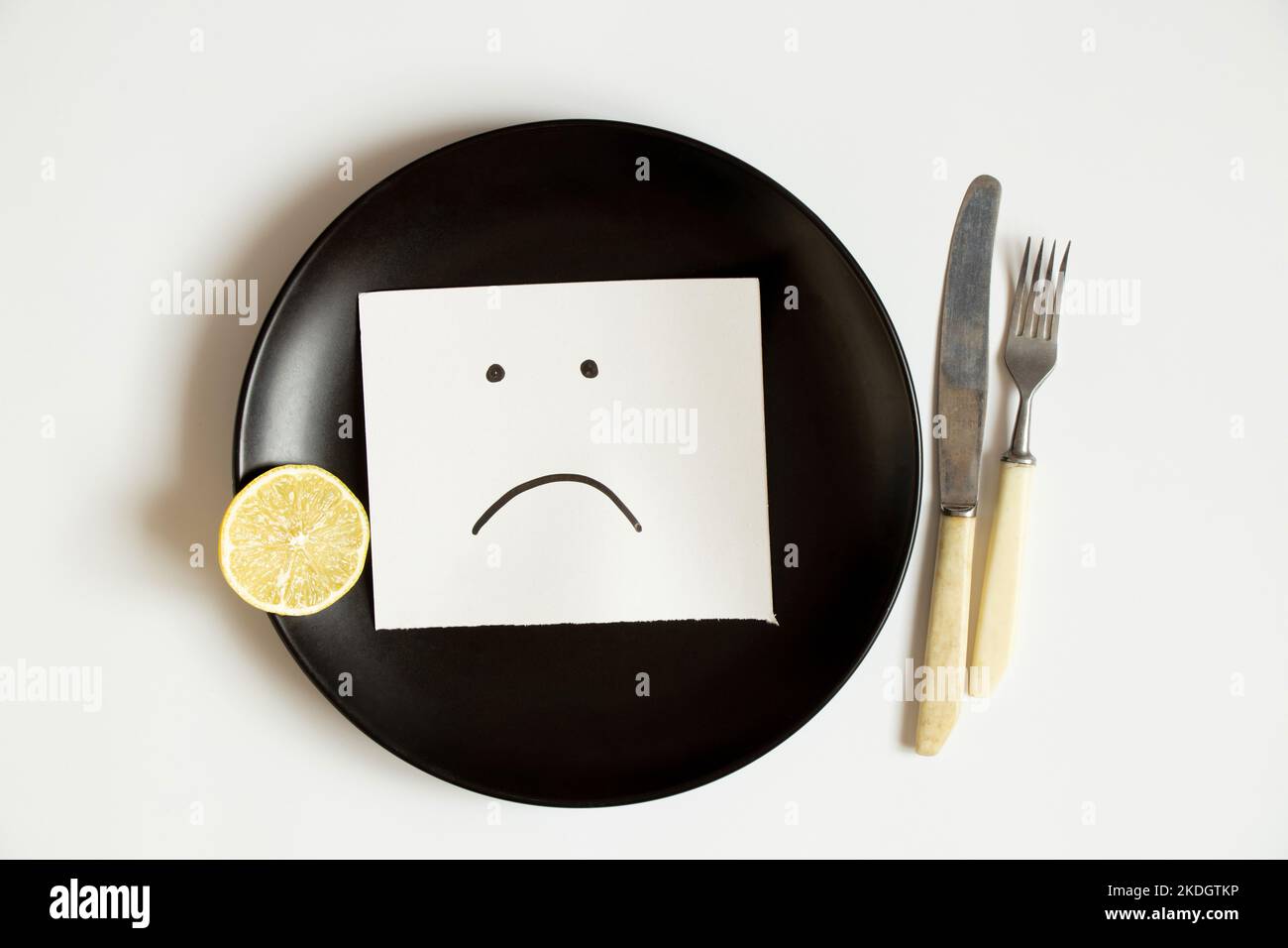 Ein trauriges Emoticon wird auf Papier gezeichnet, liegt auf einem Teller auf weißem Hintergrund, Traurigkeit Traurigkeit und kein Essen Stockfoto