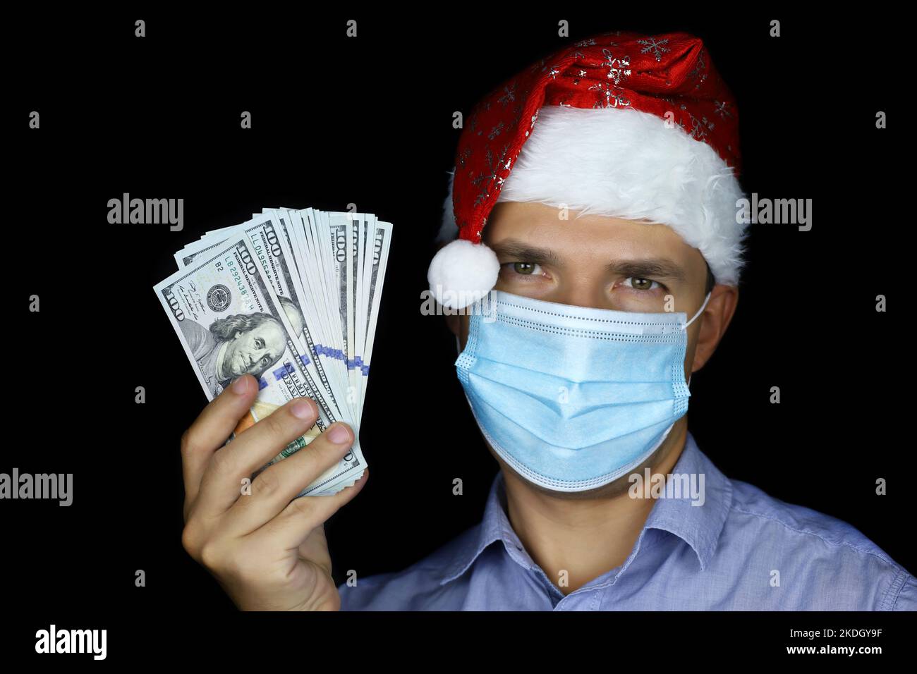 Porträt eines Mannes in Maske und Weihnachtsmütze mit US-Dollar in der Hand auf schwarzem Hintergrund. Bonusgeschenk für Weihnachtsfeier, Neujahr Stockfoto