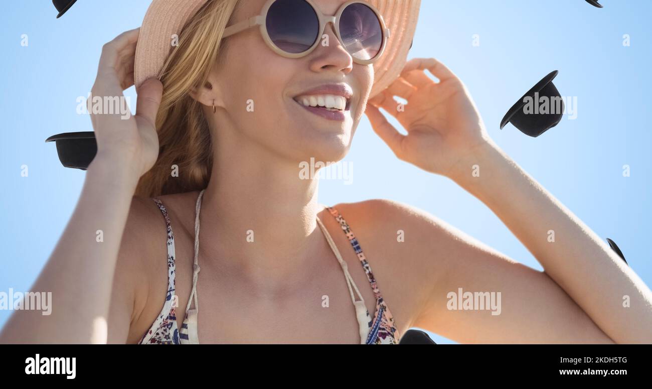Fröhliche junge weiße Frau mit Sonnenbrille und Hut und digitaler Kopfbedeckung auf blauem Hintergrund Stockfoto