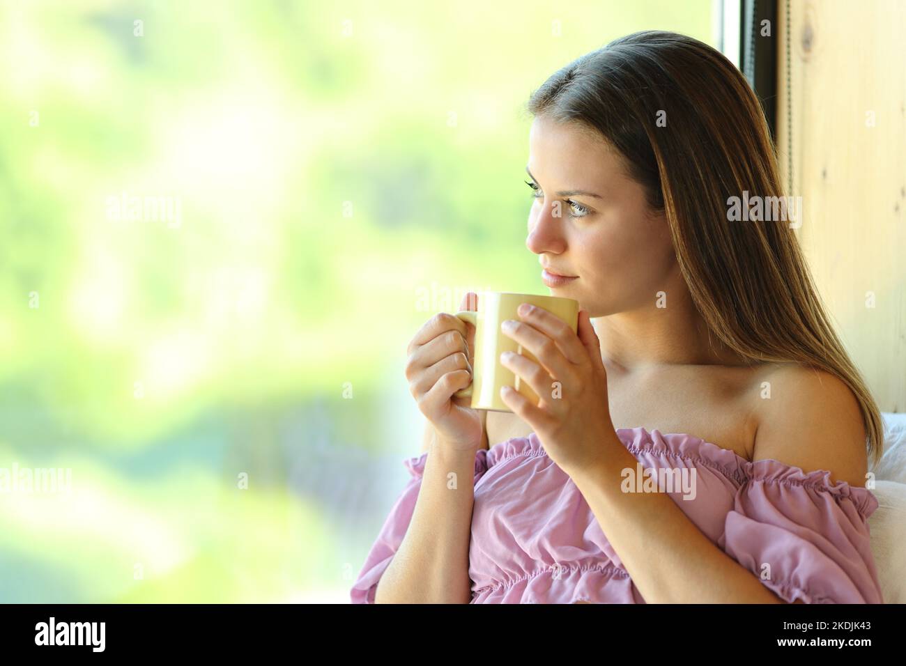 Fröhlicher Teenager, der Kaffeebecher hält und durch ein Fenster zu Hause oder in einem ländlichen Hotel schaut Stockfoto