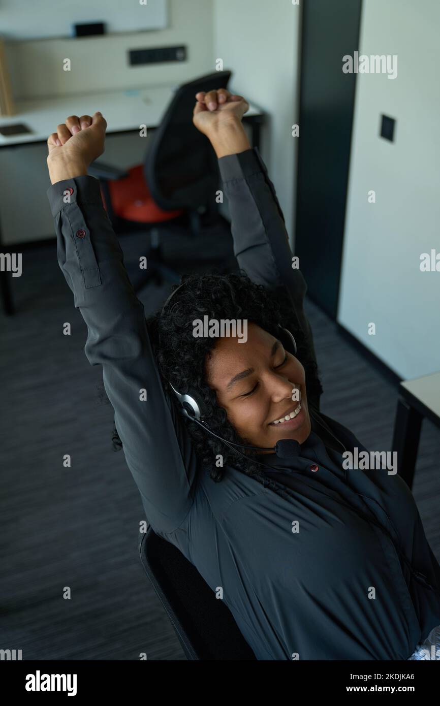 Lächelnde, multirassische Frau mit einem Mobiltelefon-Headset, das sich am Arbeitsplatz sanft streckt Stockfoto