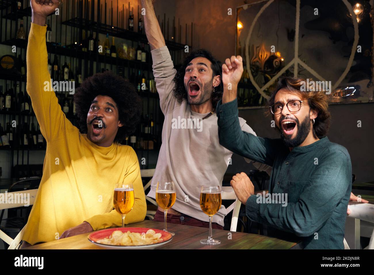 Multirassische Gruppe von männlichen Freunden, die Fußballspiele an der Bar anschauen. Feiern Sie eine Partitur mit den Fäusten Stockfoto