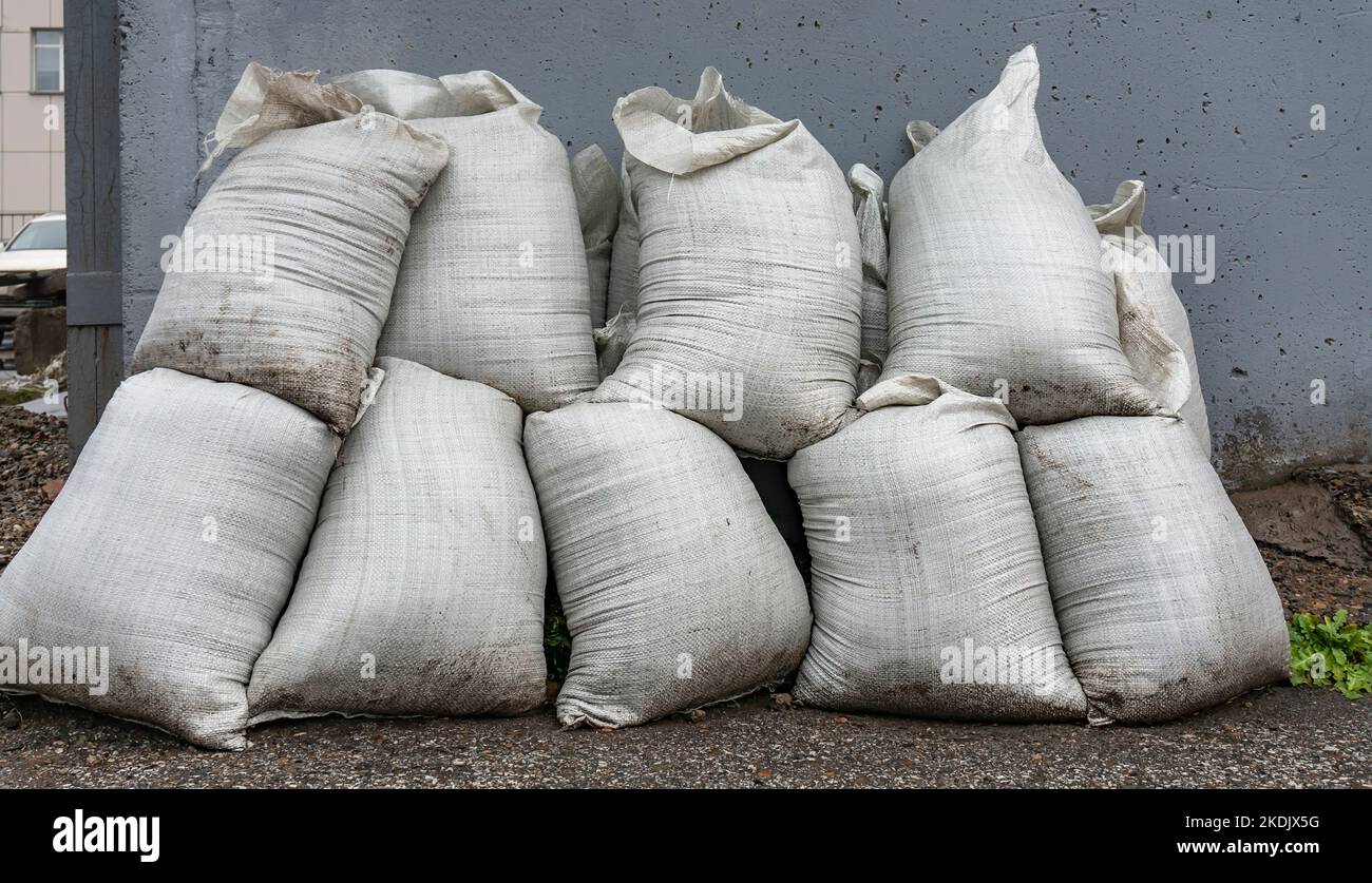 Blick auf einen Haufen weißer Säcke, die auf nassem Asphalt auf der Straße gegen die Wand eines städtischen Gebäudes öffentlicher Versorgungsunternehmen stehen Stockfoto