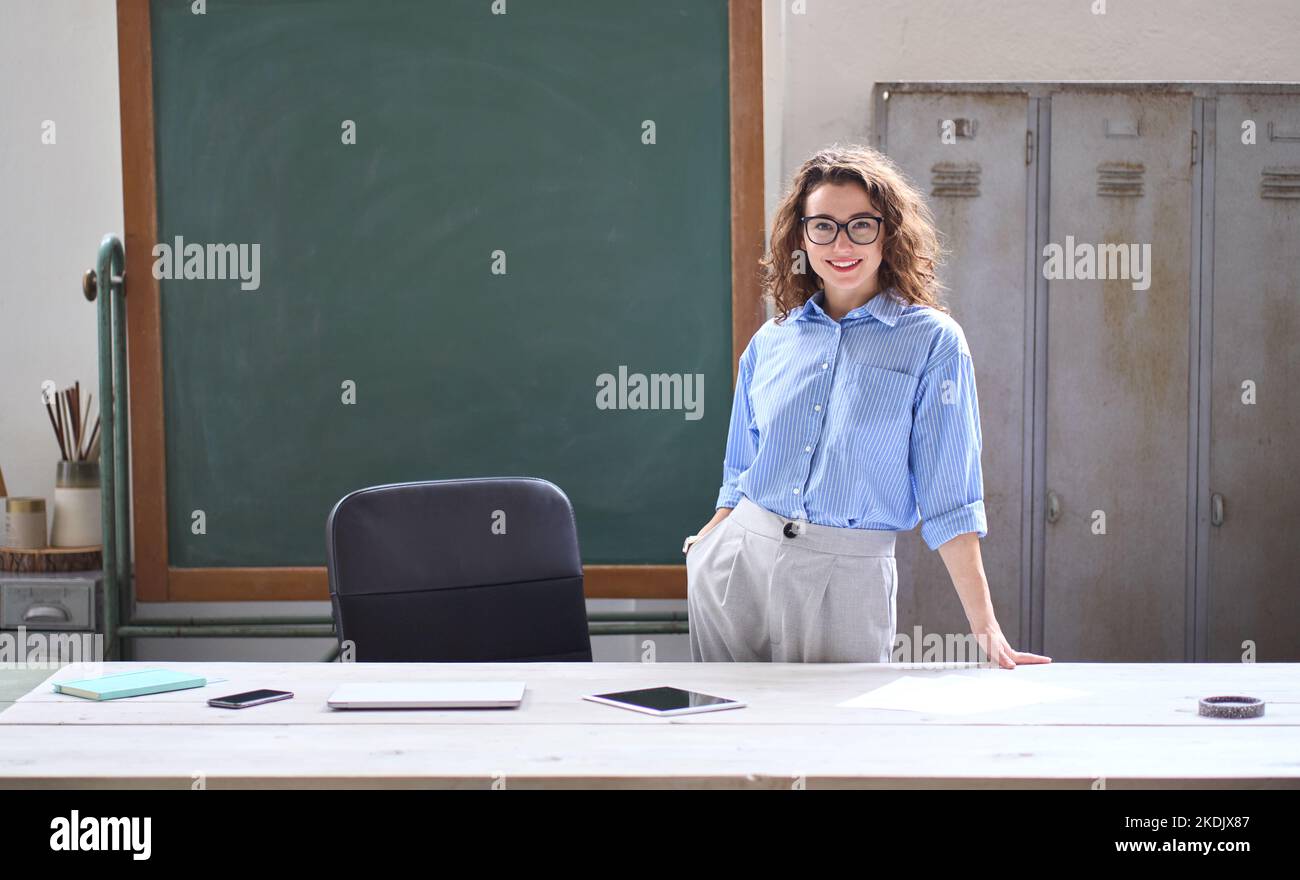 Junge Lehrerin an der Schule, die im Klassenzimmer vor der Tafel am Schreibtisch steht. Stockfoto