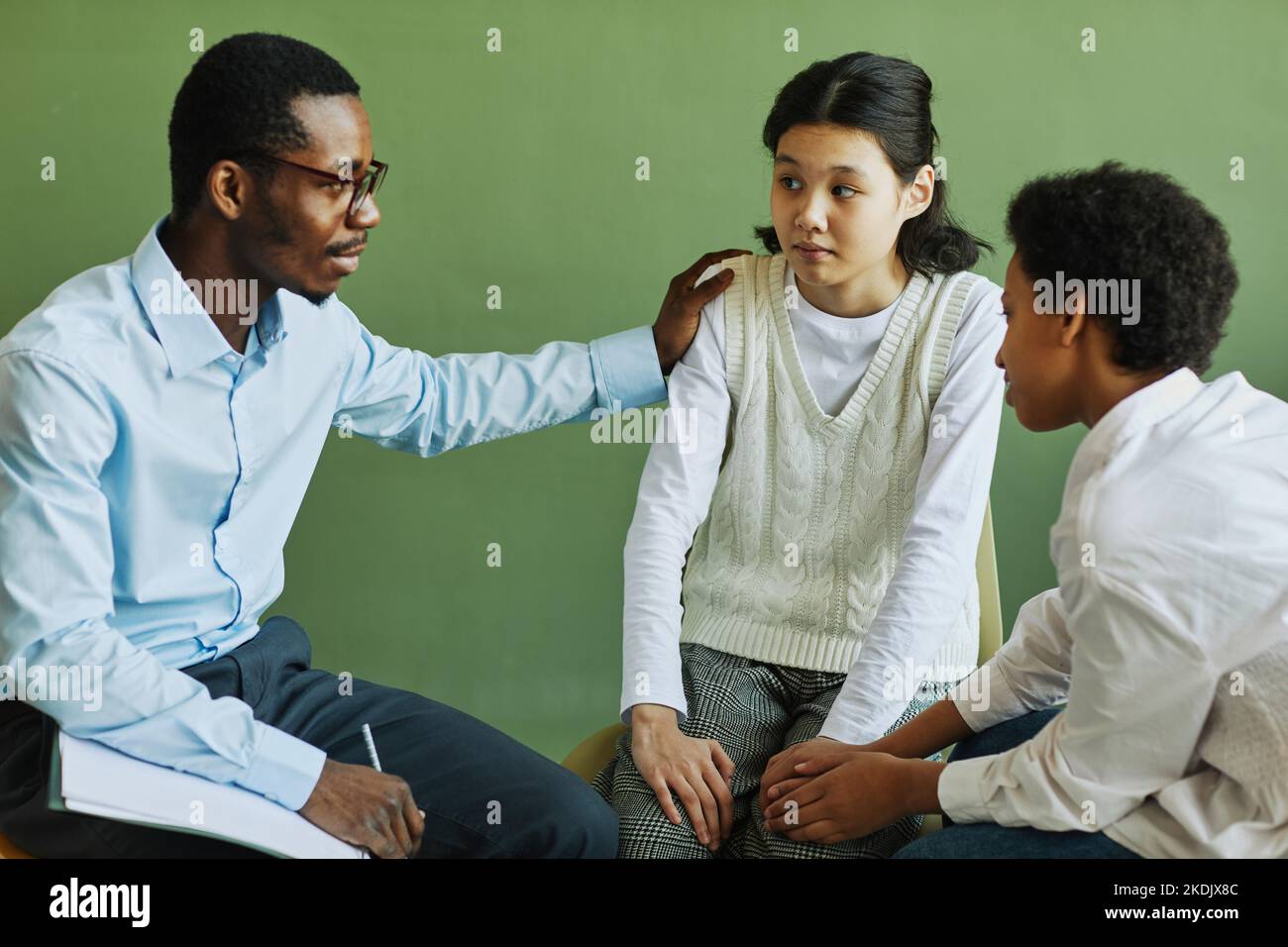 Der junge afroamerikanische Psychologe unterstützt das asiatische Schulmädchen und gibt bei der Sitzung Ratschläge, während er die Hand auf ihrer Schulter hält Stockfoto