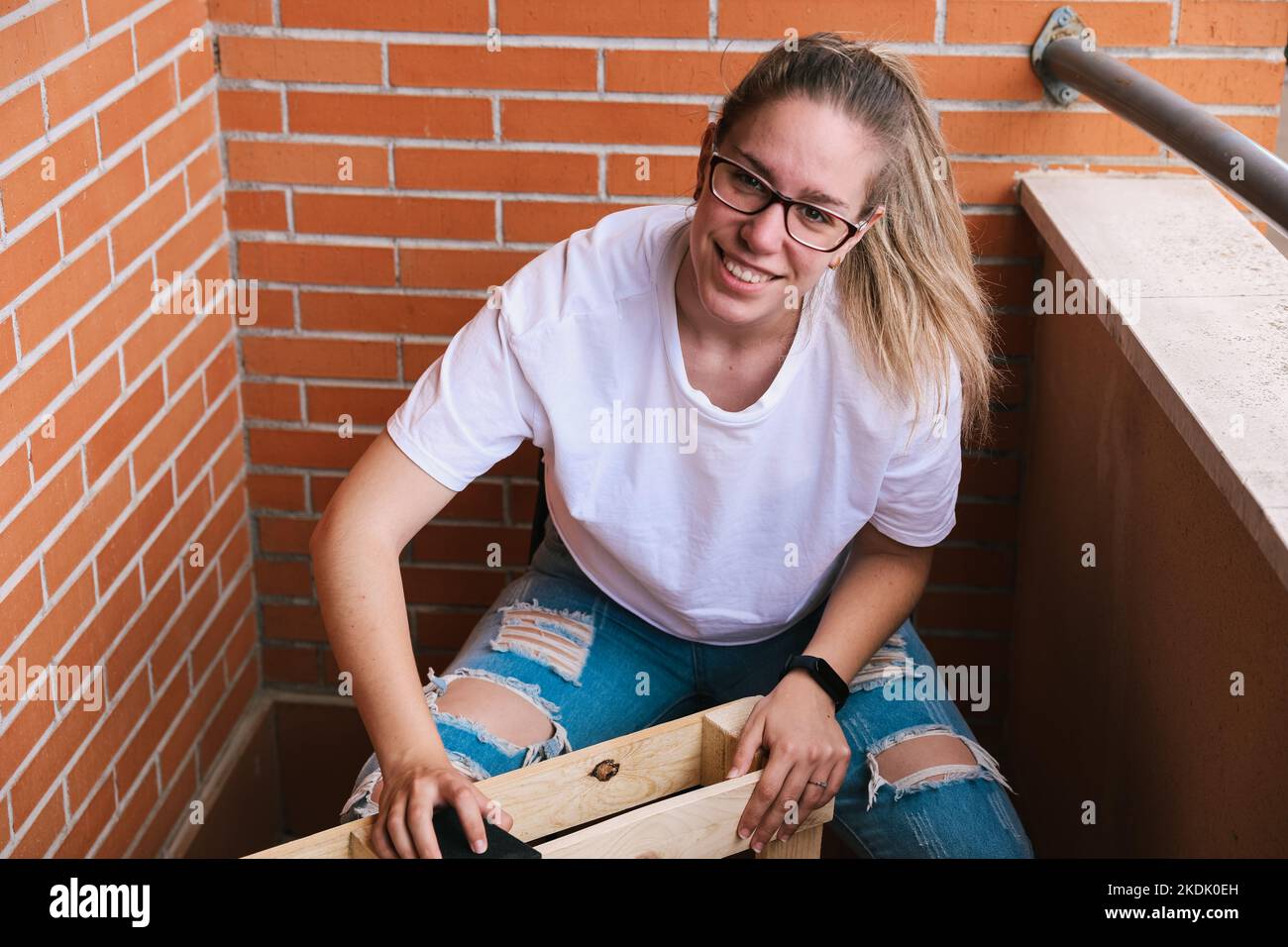 Nahaufnahme einer blonden Frau mit einer lächelnden Brille, die mit einem Sandpapier eine Palette auf der Terrasse angeschliffen hat. Renovierung des Hauses Stockfoto