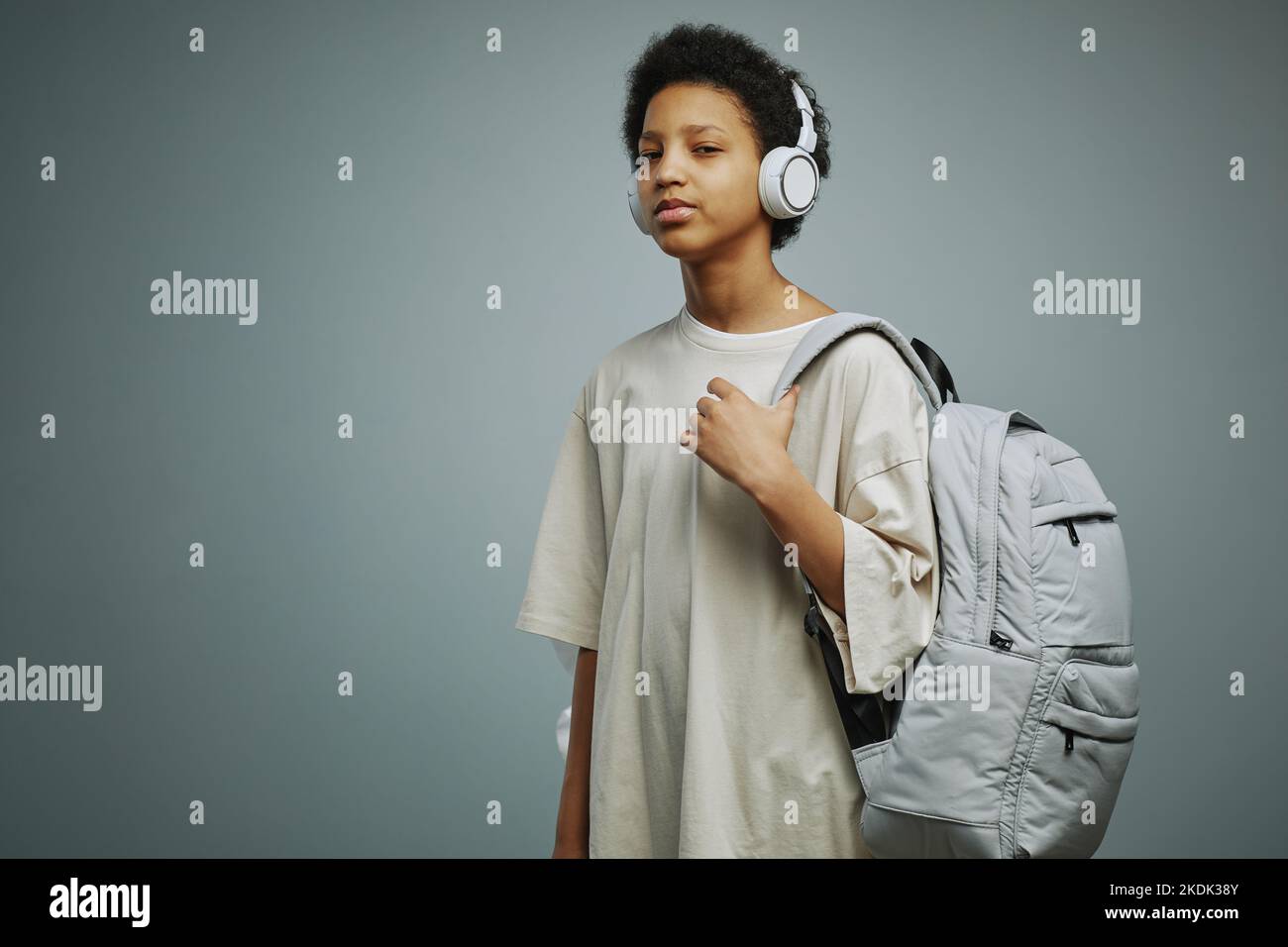 Niedliches afroamerikanisches Schulmädchen mit Rucksack auf der Schulter und Kopfhörern am Kopf, das Musik hört und die Kamera anschaut Stockfoto