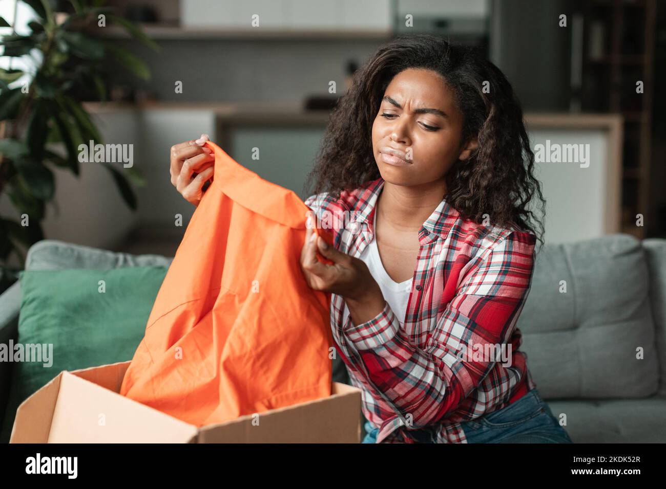 Traurige unzufriedene schwarze junge Frau in lässigen Anblicken auf Kleidung, packt Paket im Wohnzimmer Interieur aus Stockfoto