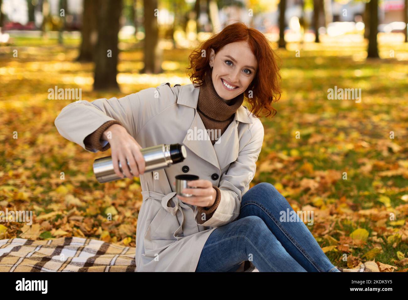 Lächelnd sitzt die hübsche kaukasische Millennials-Dame mit roten Haaren im Regenmantel auf kariert und gießt heißes Getränk aus Thermoskanne Stockfoto