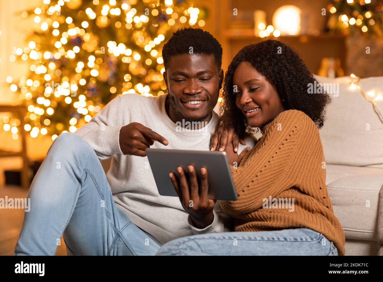 Lächelnder junger afroamerikanischer Mann zeigt der Dame ein Tablet, führt einen Videoanruf durch, genießt den Urlaub im Wohnzimmer Stockfoto