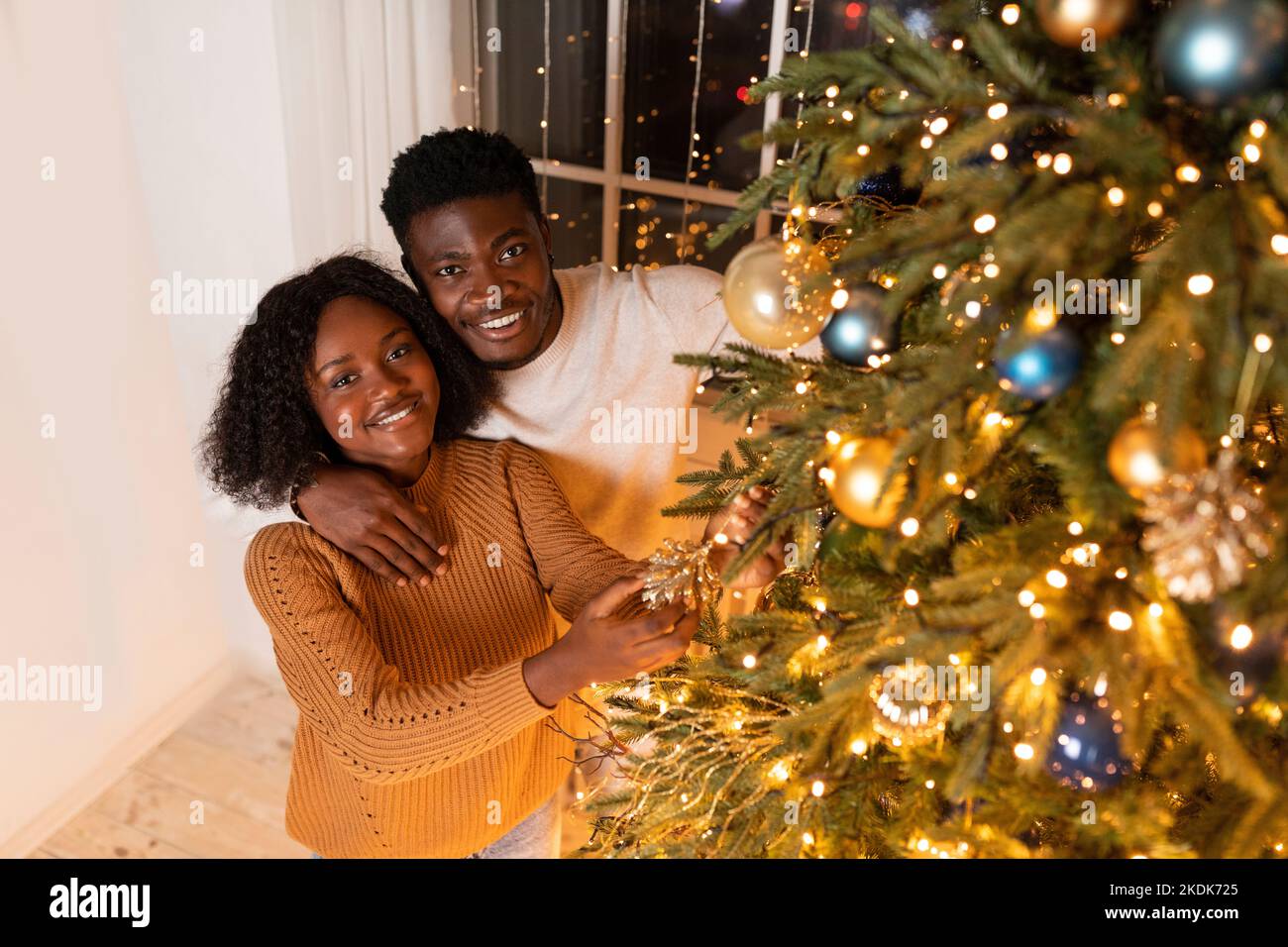 Lächelnde, tausendjährige schwarze Frau und Ehemann schmücken den Weihnachtsbaum mit Lichtern und Spielzeug im gemütlichen Wohnzimmer Stockfoto
