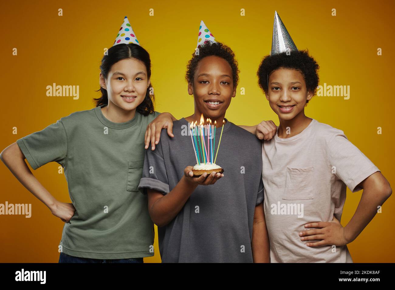 Drei Jugendliche multikulturelle Freunde in Geburtstagsmützen und Casualwear stehen vor der Kamera, während Junge Kuchen mit Kerzen hält Stockfoto
