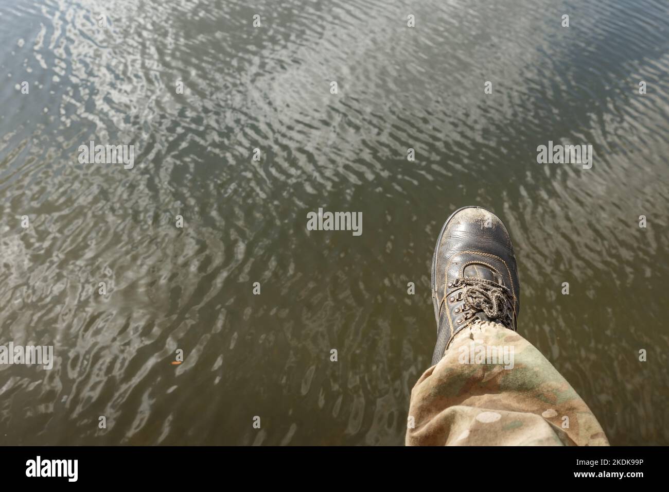 Niedriger Personenabschnitt über Wasser. Das rechte Bein eines Mannes in Camouflage-Hosen und alten Stiefeln baumelt über dem Wasser. Stockfoto
