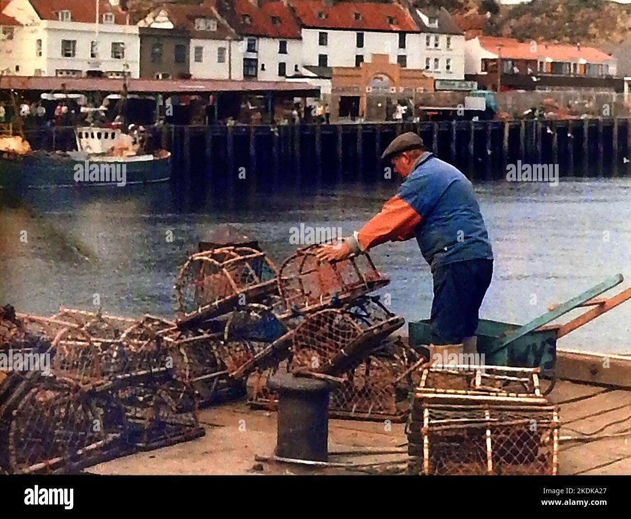 Eine alte Momentaufnahme eines Fischers aus Whitby, Yorkshire, der traditionelle handgemachte Krabben-/Hummertöpfe stapelt und in seiner selbstgemachten Schubkarre transportiert wird. Der Pier auf der Westseite des Flusses Esk, mit seinem Fischmarkt Gebäude kann mit einem lokalen Fischerboot in der Nähe vor Anker gesehen werden. Stockfoto