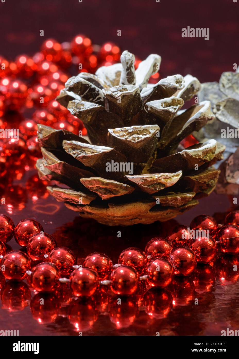 Weihnachtsdekoration mit einem Tannenzapfen und einer roten Perlengirlande auf einem rot glänzenden Hintergrund. Stockfoto