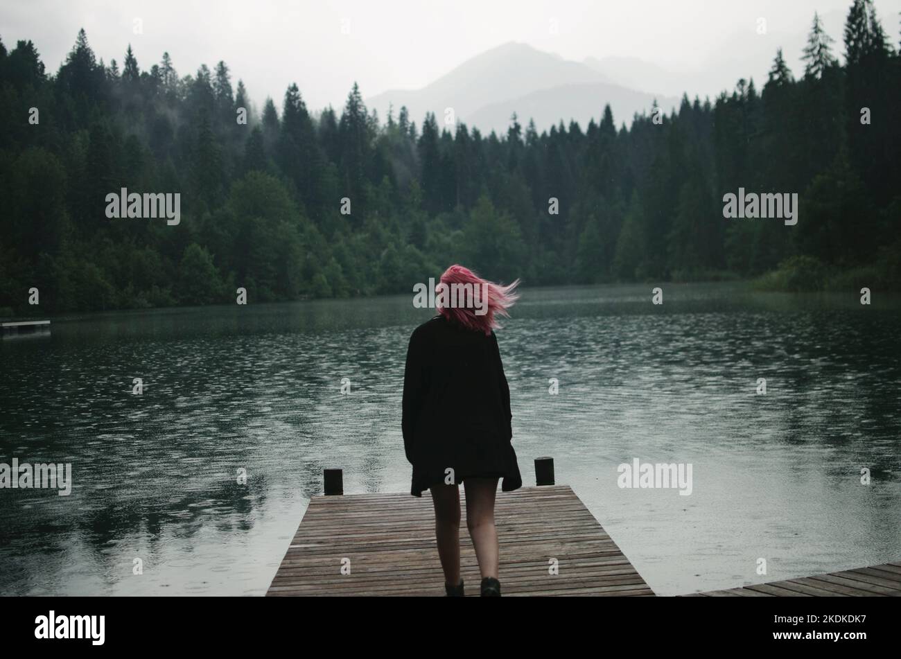 Frau mit rosafarbenem Haar, die bei Regen einen Steg am Crestasee in der Schweiz hinunterläuft Stockfoto