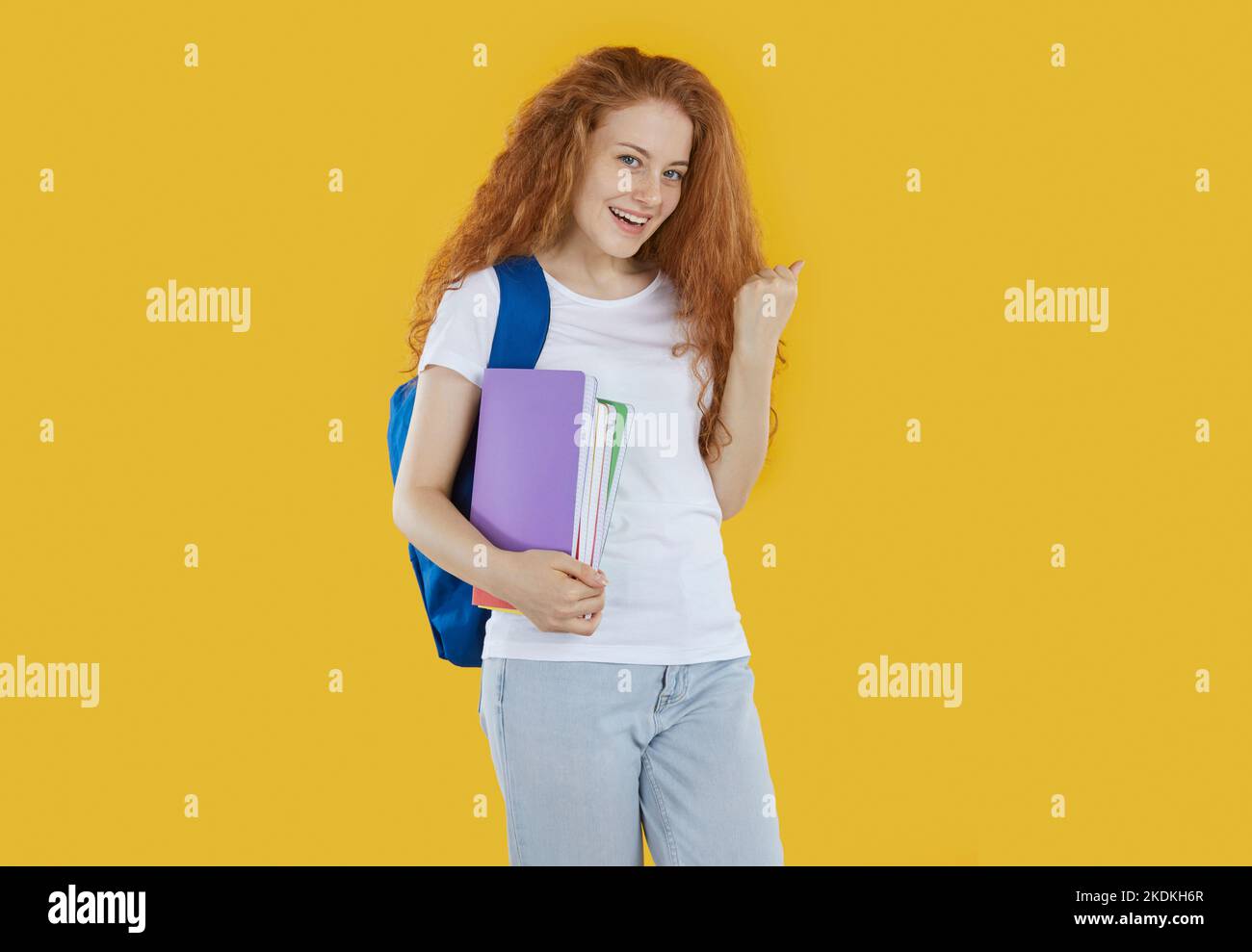 Happy Rotschopf Mädchen Studentin mit Rucksack und Copybooks zeigt ja Geste auf gelbem Hintergrund. Stockfoto