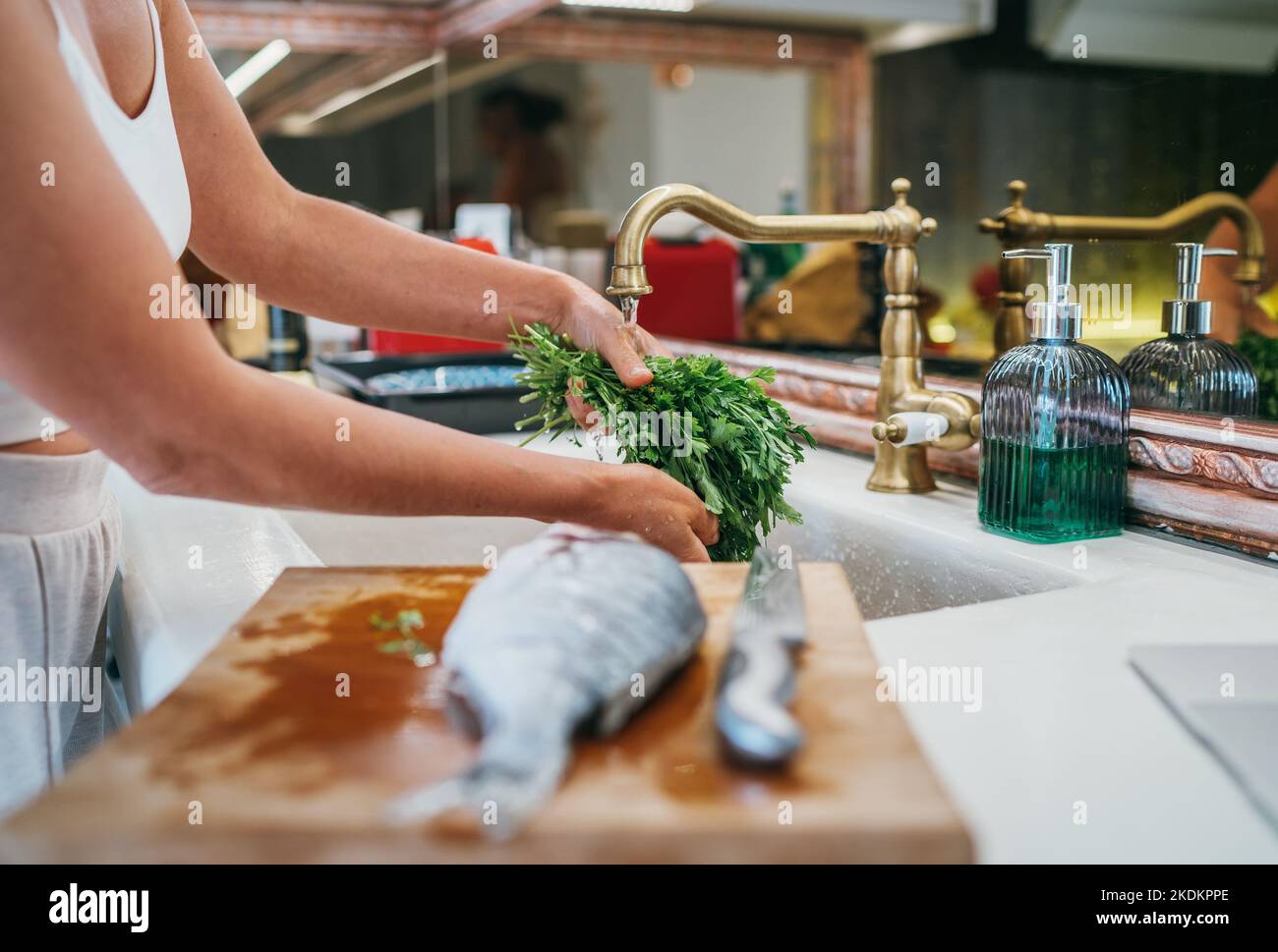 Waschen der frischen grünen Petersilie Kraut unter dem Spülbecken Wasserhahn und Fisch auf Schneidebrett Vorbereitung zum Kochen. Gesunde Lebensmittel und frische Meeresfrüchte Stockfoto
