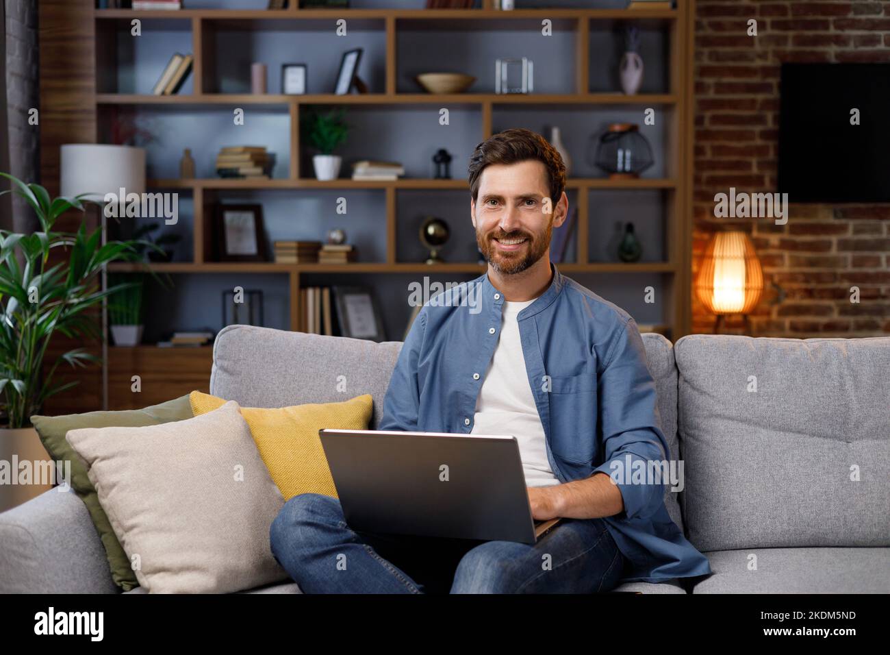 Ein hübscher Mann, der in einer stilvollen Wohnung auf dem Sofa sitzt und am Laptop arbeitet. Arbeiten im Internet, Nutzung sozialer Netzwerke, Unterhaltung in der Wohnung Stockfoto