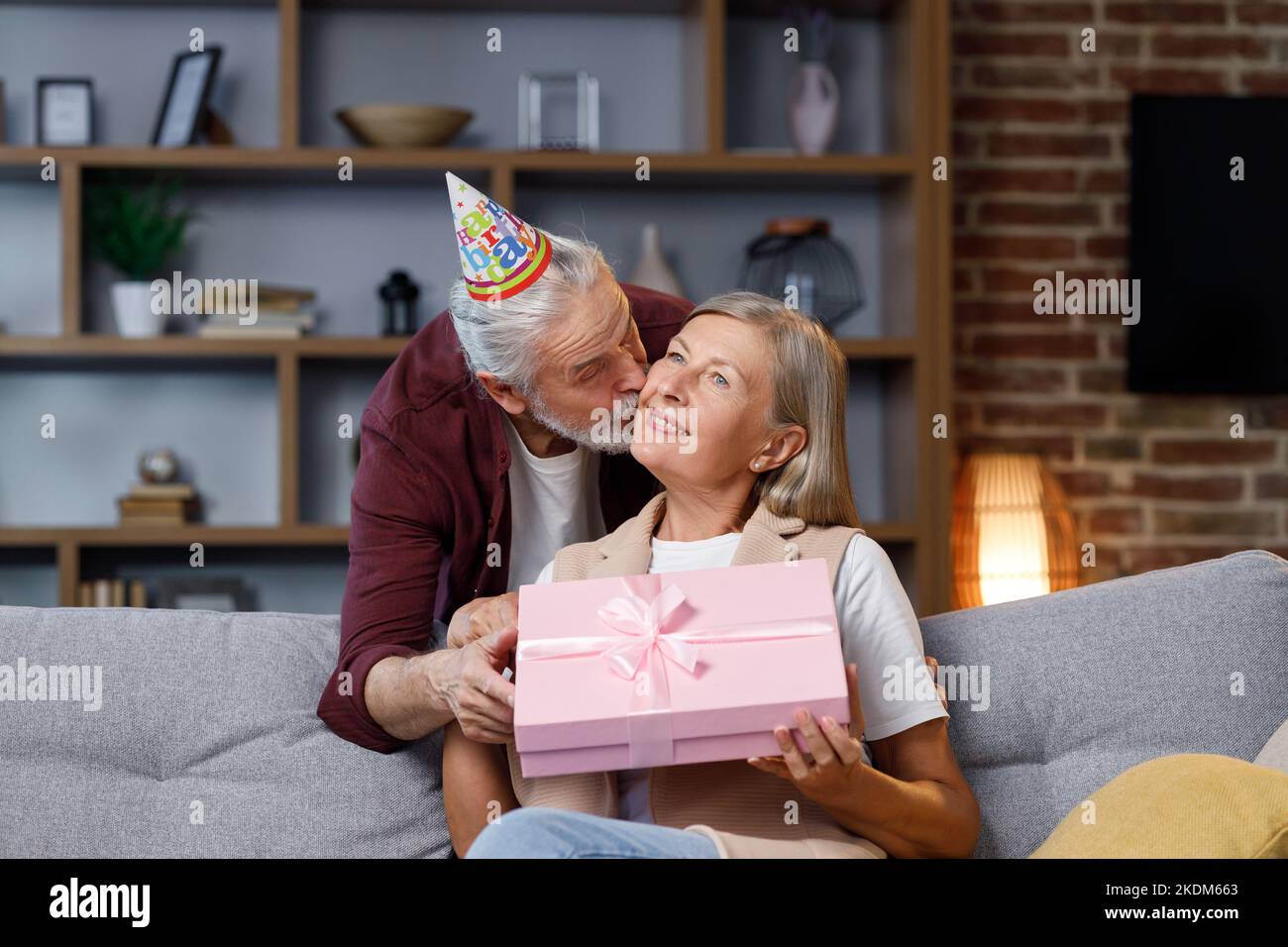 Fürsorglicher alter Ehemann, der aufgeregt reife Frau ein romantisches Geburtstagsgeschenk gibt, liebevoller älterer Mann, eine glückliche Frau mittleren Alters, eine rosa Schachtel, einen Kuss Stockfoto