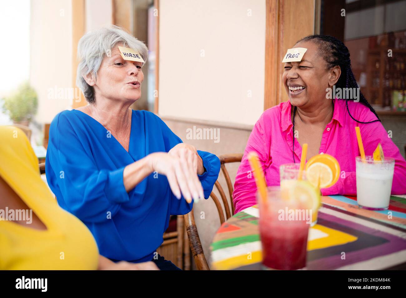 Multirassische ältere Freunde, die Spaß beim Spielen von „Guess Who Stirnaufklebern“ im Bar-Restaurant haben - Fokus auf afrikanisches Frauengesicht Stockfoto