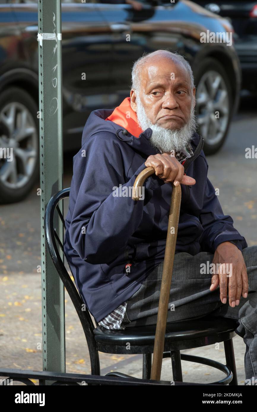Ein älterer Mann, wahrscheinlich Muslim, sitzt und lehnt sich auf seinen Stock. An der 74. Street in Jackson Heights, Queens, New York City. Stockfoto