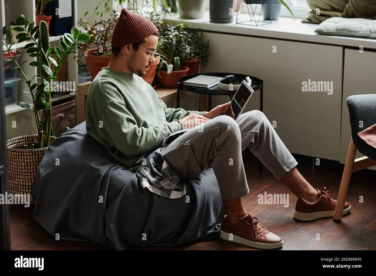 Junger seriöser Webdesigner mit Laptop, der sich auf das Netzwerk konzentriert, während er in einem bequemen Sessel im Büro mit vielen grünen Pflanzen sitzt Stockfoto