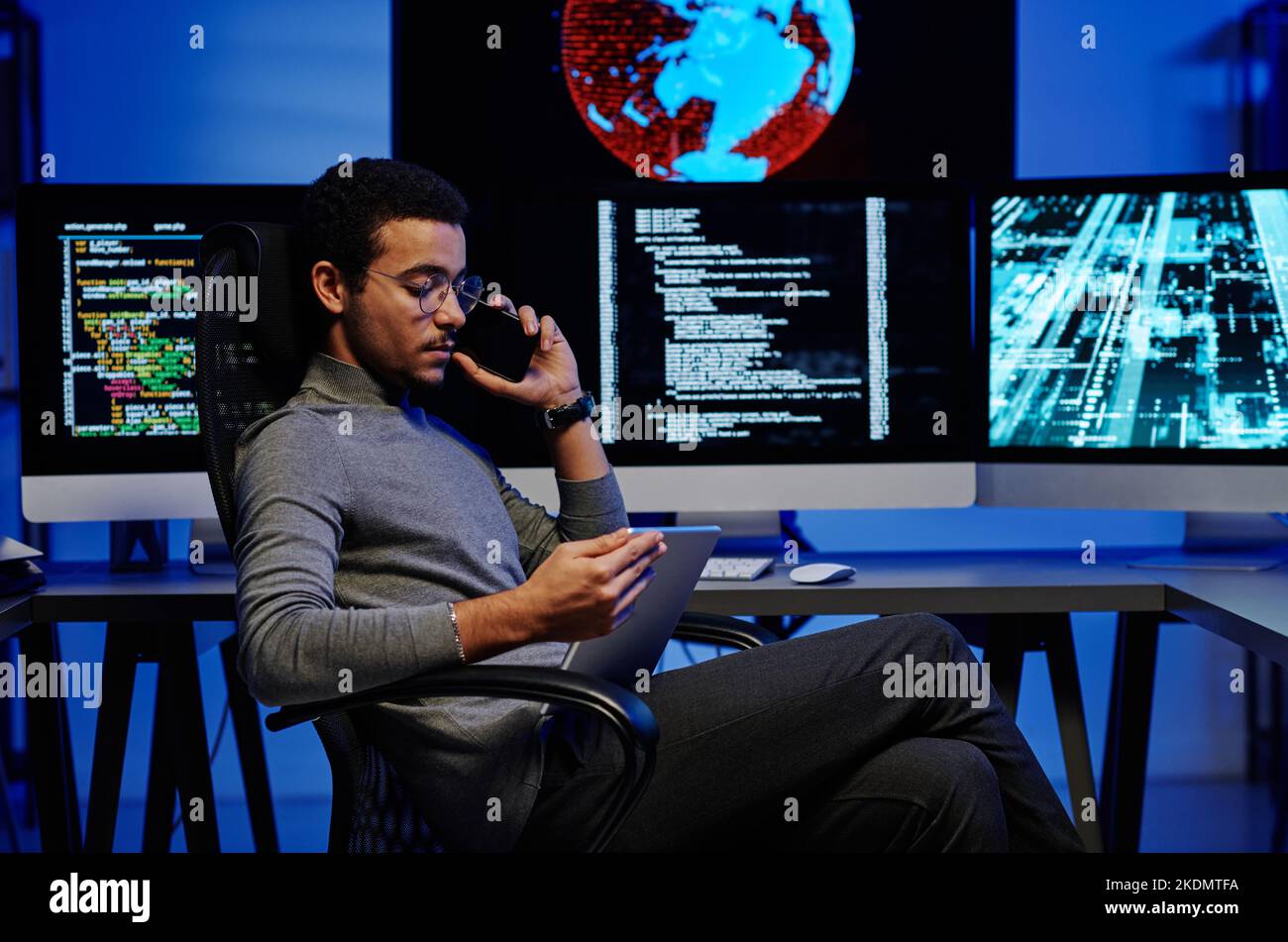 Seitenansicht des jungen beschäftigten männlichen Programmierers, der auf dem Smartphone spricht und Online-Informationen oder Videos auf dem Tablet-Bildschirm durchschaut Stockfoto