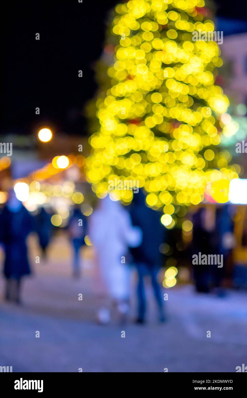 Unscharfer Hintergrund. Weihnachtsbaum, verzierte leuchtende Lichter, Gebäude, verschwommene Silhouetten von Menschen, die in der Winternacht auf dem Stadtplatz herumlaufen. Schönes Neujahr und Weihnachten verschwommen Hintergrund Stockfoto