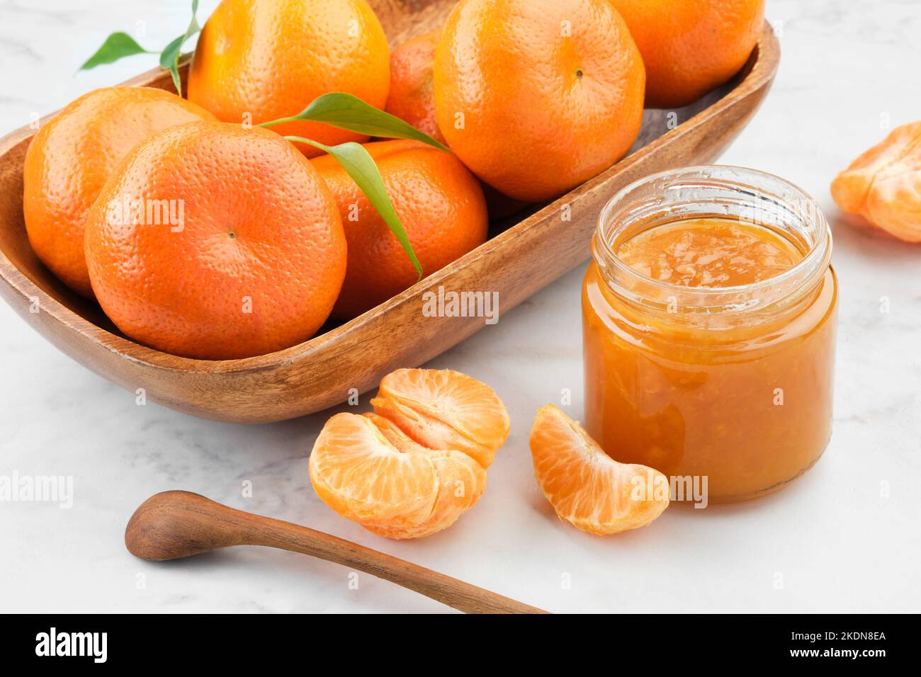 Krug mit gesunder Mandarinenkonfitüre und Holzschüssel mit Mandarinenorangen-Früchten auf weißem Küchentisch. Stockfoto