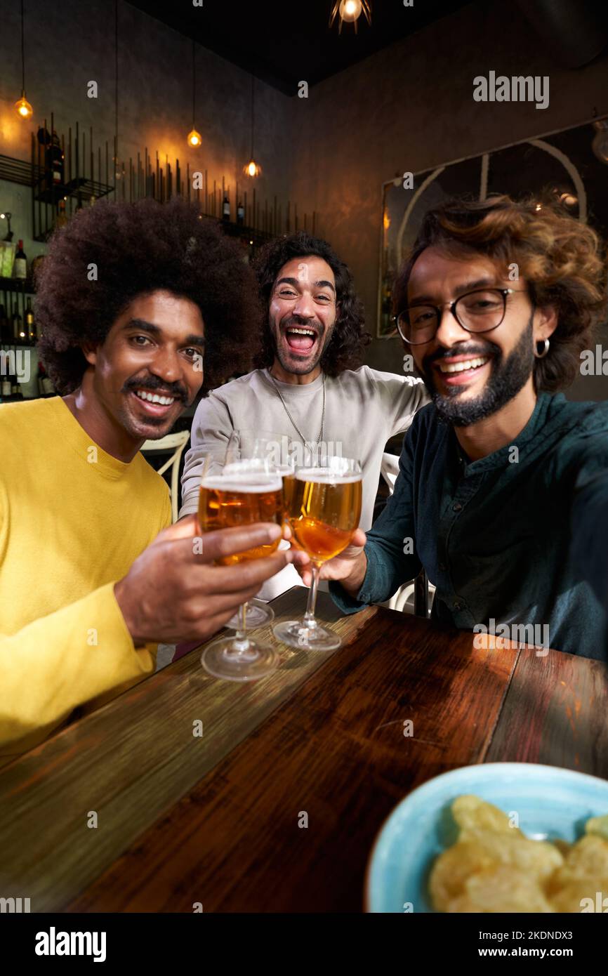 Vertikales Selfie von drei männlichen Freunden, die fröhlich in einer Bar toasten und mit großem Lächeln auf die Kamera blicken. Stockfoto