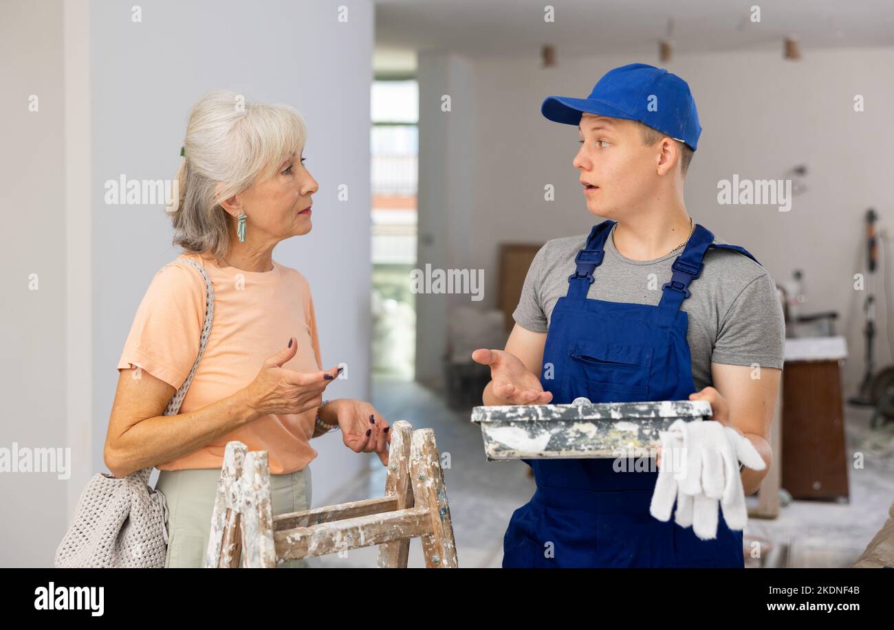 Ein junger Baumeister in blauen Overalls und eine ältere Frau diskutieren über die Renovierung eines Zimmers in einer Wohnung Stockfoto