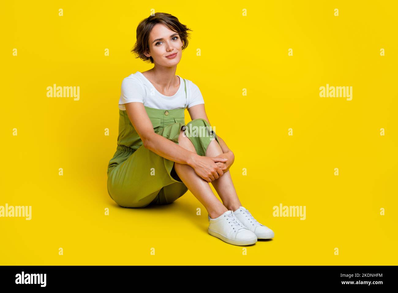 In voller Länge Foto von charmanten jungen Frau sitzen Boden Umarmung Knie Shopping promo tragen trendige khaki Kleidungsstück auf gelbem Hintergrund isoliert Stockfoto