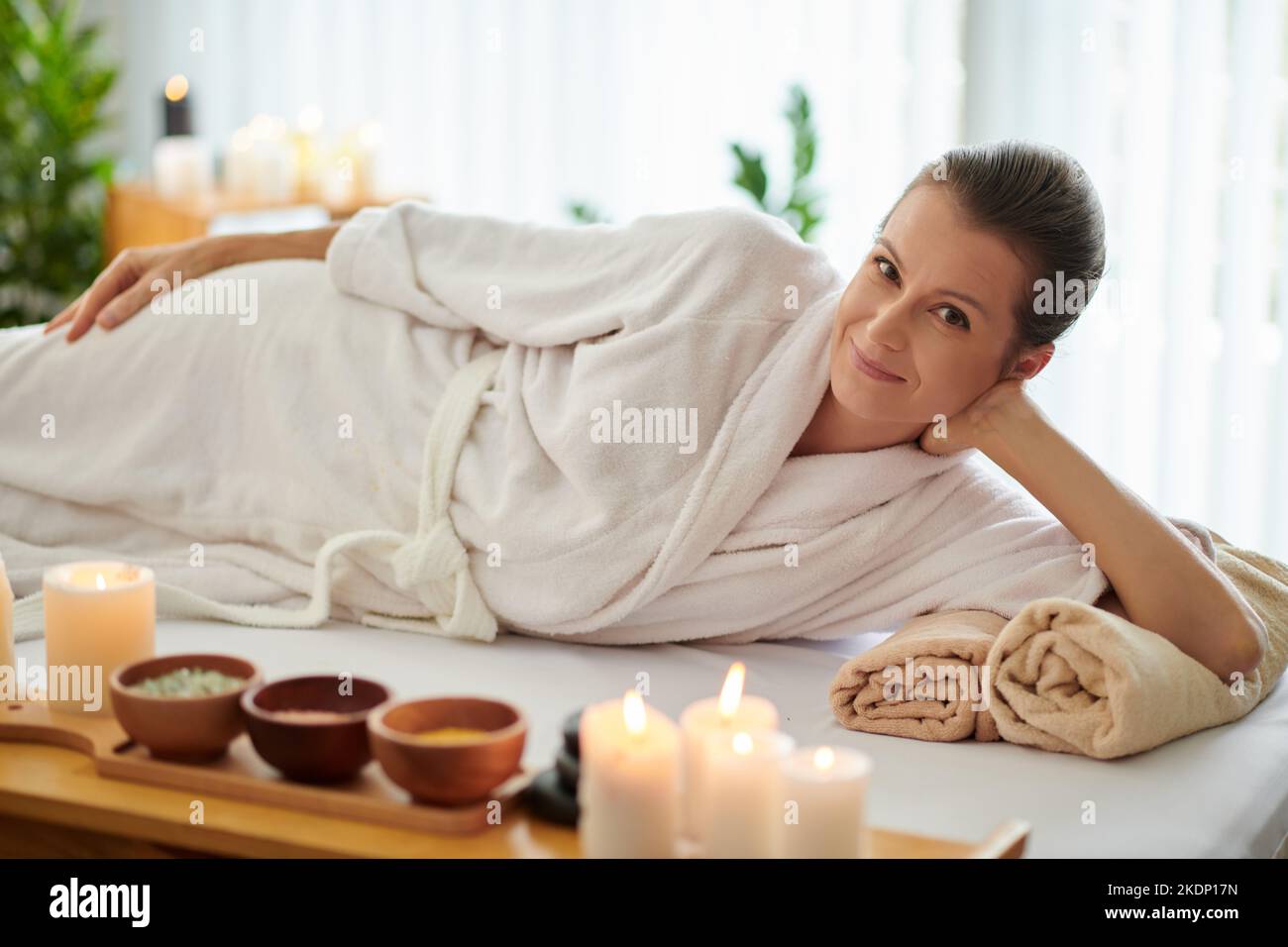 Lächelnde Frau im Bademantel, die sich nach einer Massage im Spa-Salon auf dem Bett entspannt hat Stockfoto