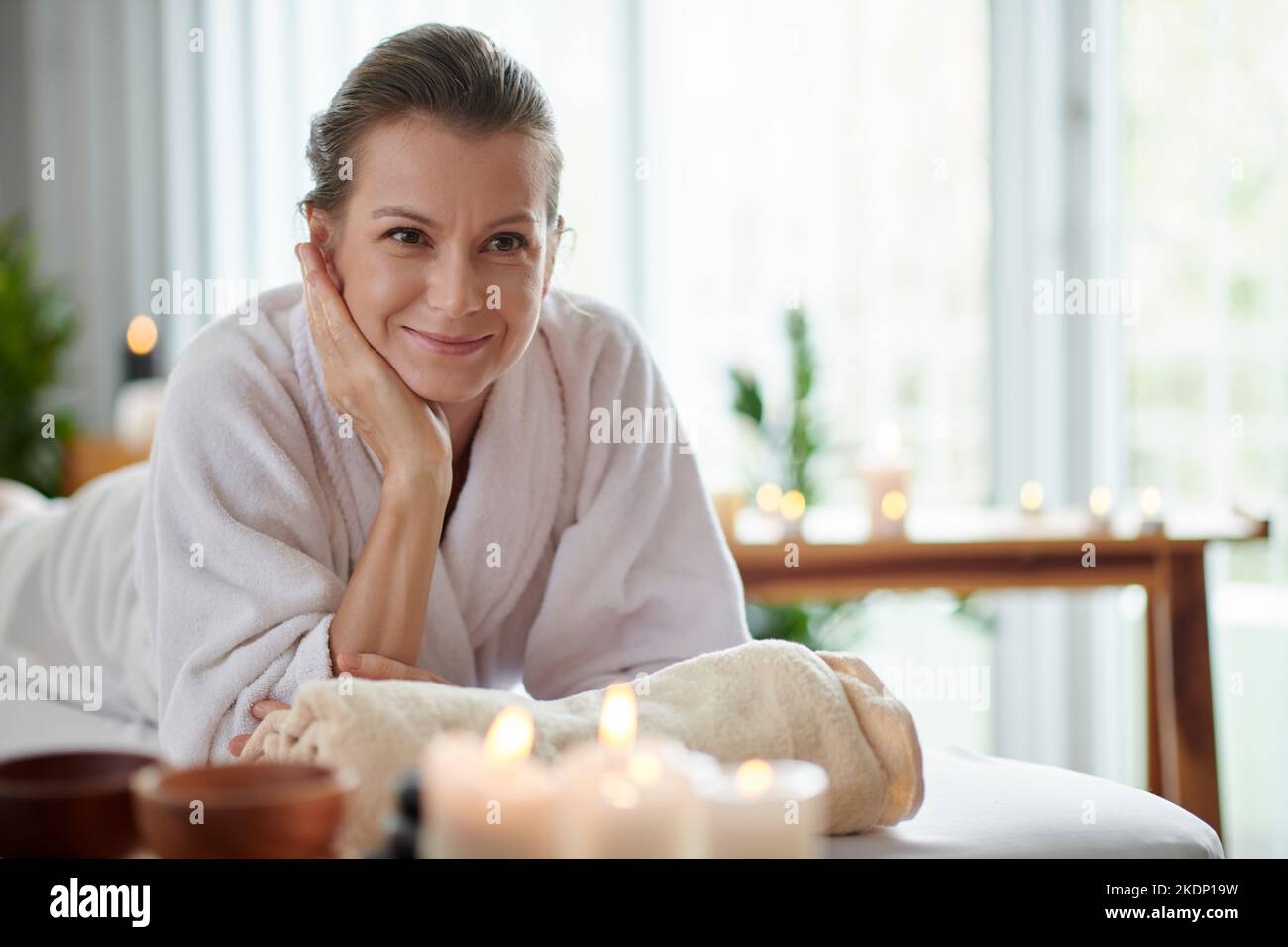 Eine glückliche Frau mittleren Alters im weichen Bademantel, die sich im Spa-Salon mit brennenden Kerzen entspannen kann Stockfoto