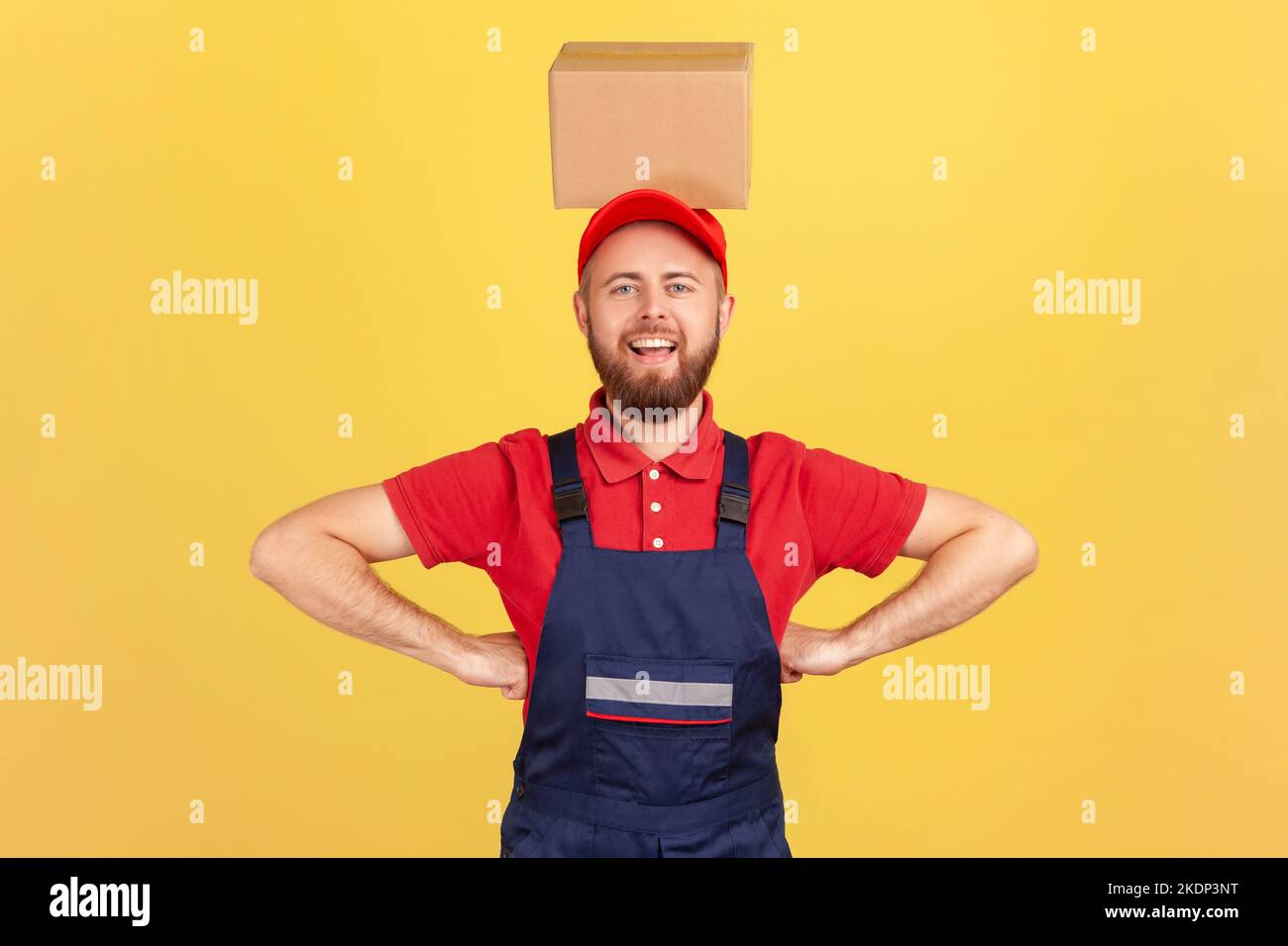 Porträt eines lustigen Kuriermanns in blauer Uniform, der mit den Händen auf den Hüften stand und den Karton auf dem Kopf hielt und lächelte auf die Kamera. Innenaufnahme des Studios isoliert auf gelbem Hintergrund. Stockfoto