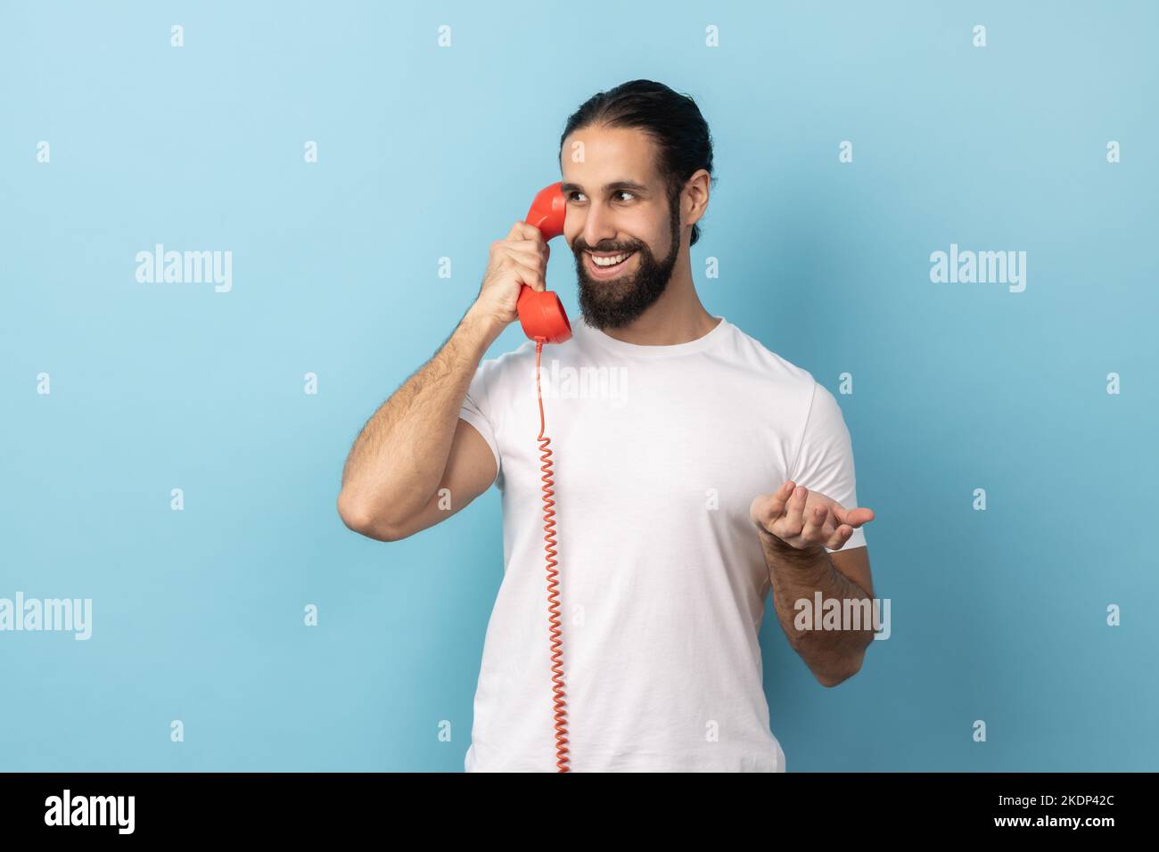 Porträt eines positiven Mannes mit Bart in weißem T-Shirt, der auf einem roten Retro-Telefon spricht, angenehme Gespräche führt, mit einem Lächeln wegschaut Innenaufnahme des Studios isoliert auf blauem Hintergrund. Stockfoto