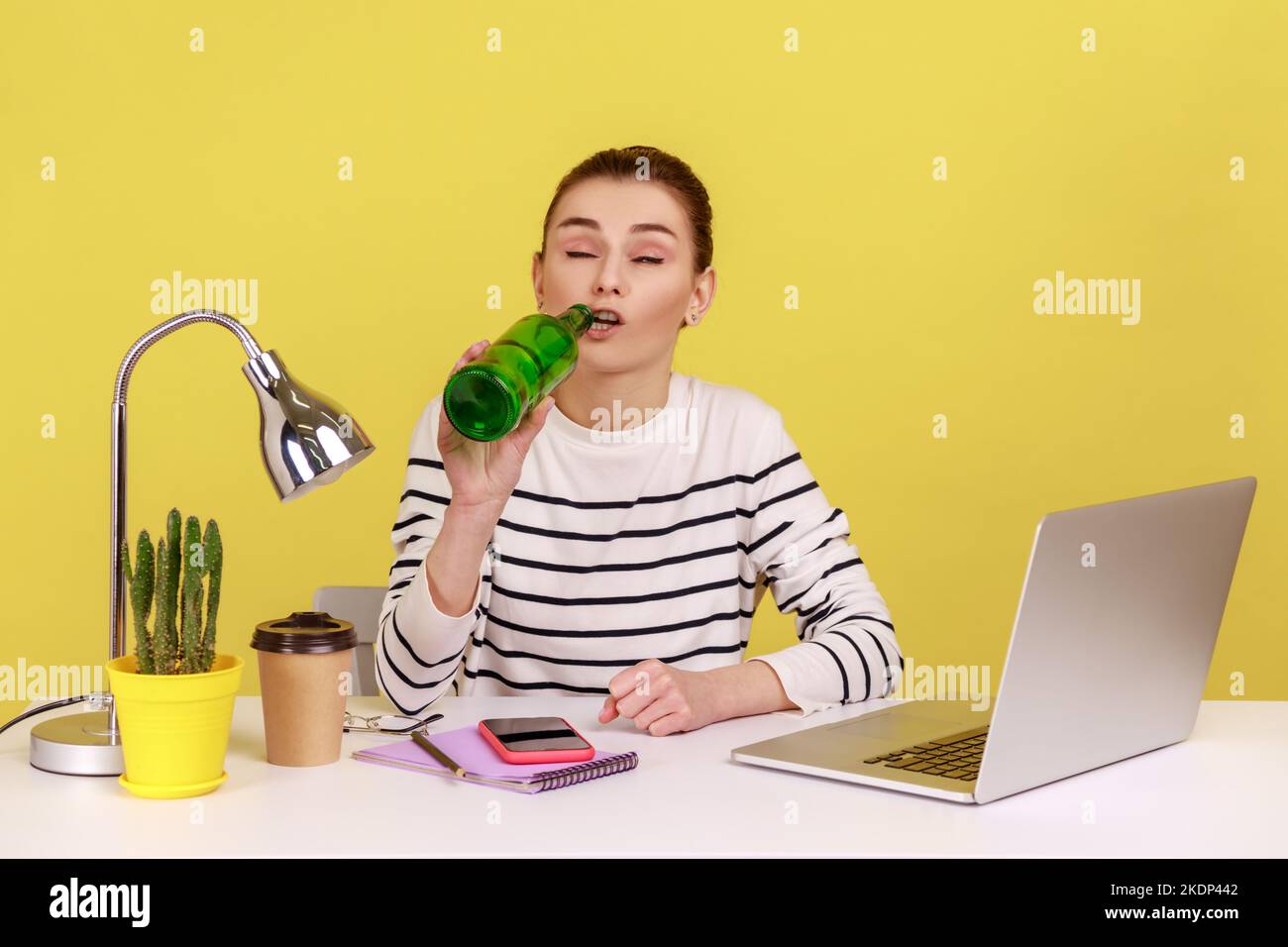 Porträt einer jungen erwachsenen Frau, die Bier trinkt, mit geschlossenen Augen am Arbeitsplatz sitzt und während des Arbeitstages betrunken ist. Studio-Innenaufnahme isoliert auf gelbem Hintergrund. Stockfoto