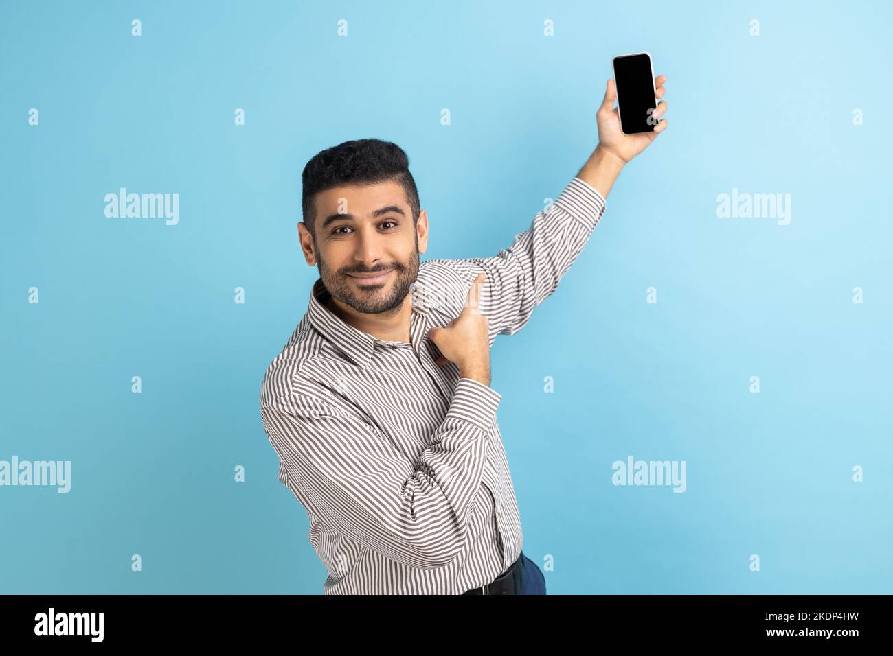 Porträt eines Geschäftsmannes, der Videoanruf unterhielt, die Kamera betrachtend, mit einem Mockup-Rohling auf das Smartphone zeigend, das gestreiftes Hemd trägt. Innenaufnahme des Studios isoliert auf blauem Hintergrund. Stockfoto