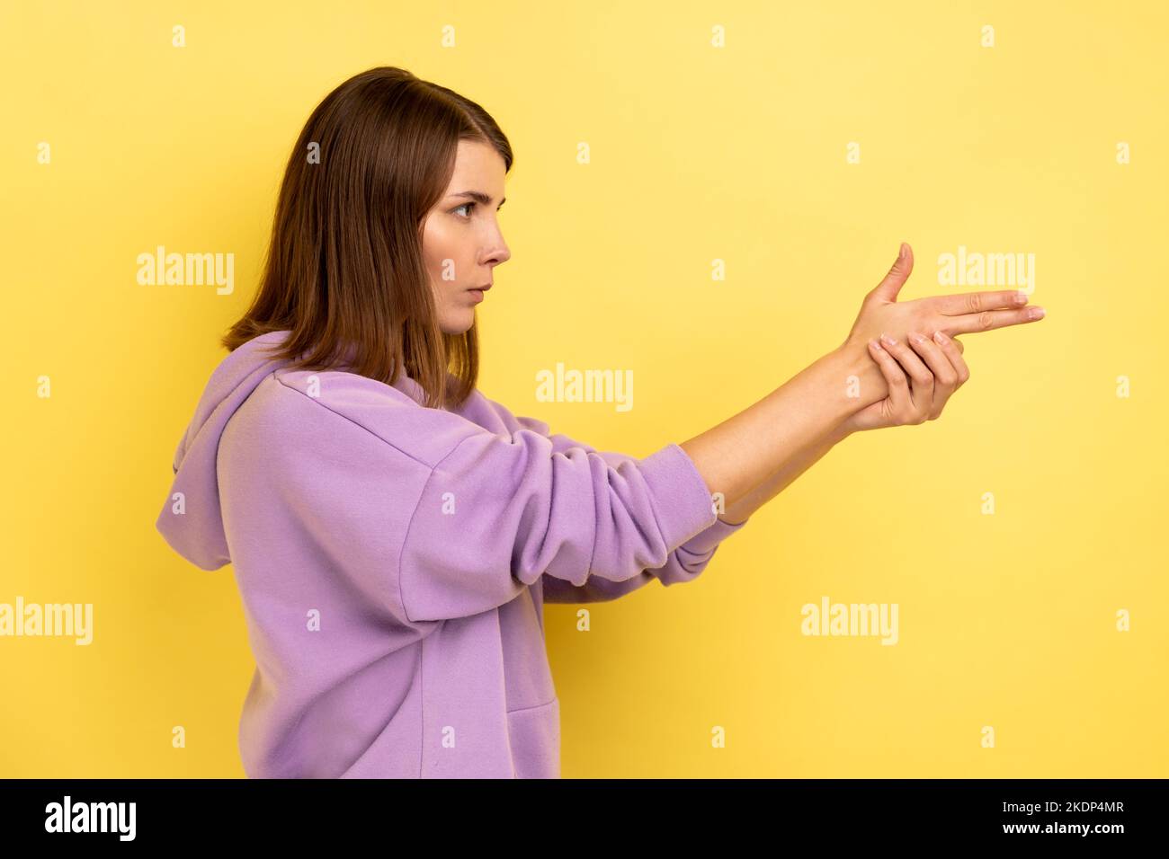 Ich werde Euch töten. Seitenansicht einer wütenden Frau, die mit den Fingergewehren auf die Kamera zeigt, mit dem Schießen droht, die Hände imitieren die Waffe und tragen einen violetten Hoodie. Innenaufnahme des Studios isoliert auf gelbem Hintergrund. Stockfoto