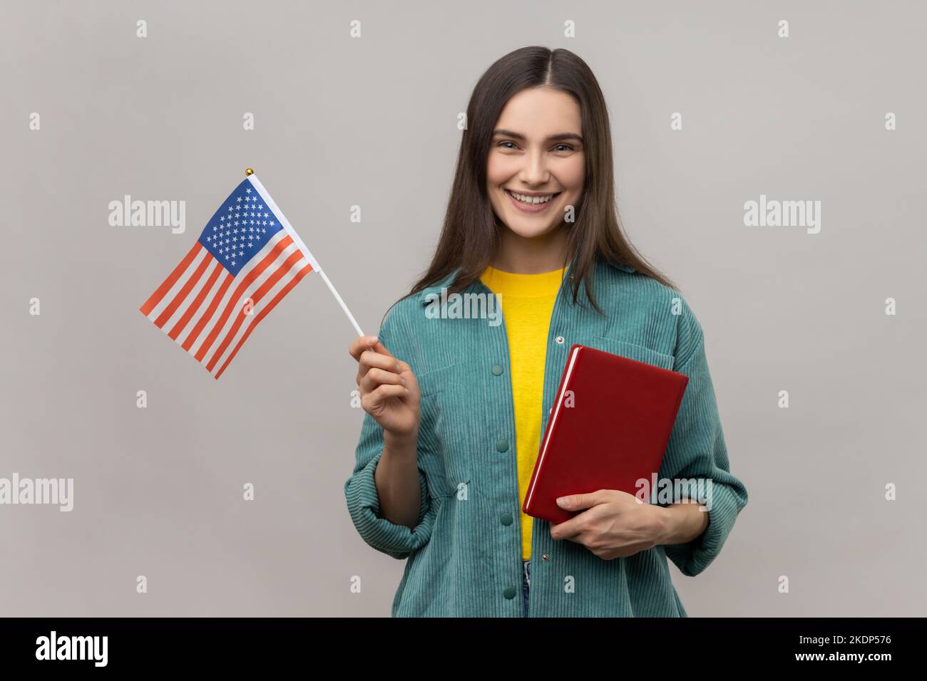 Porträt einer attraktiven, intelligent lächelnden Frau, die Buch und amerikanische Flagge in den Händen hält, Bildungskurse im Ausland, Jacke im lässigen Stil trägt. Innenaufnahme des Studios isoliert auf grauem Hintergrund. Stockfoto