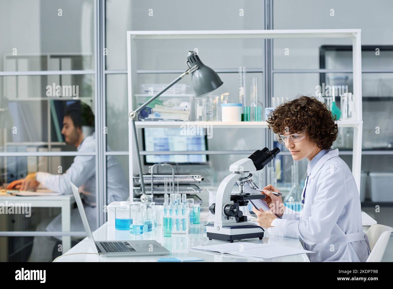 Junger Chemiker oder Mikrobiologe in Laborkittel und Schutzbrillen, die mit dem Mikroskop am Arbeitsplatz sitzen und während des Versuchs Notizen machen Stockfoto