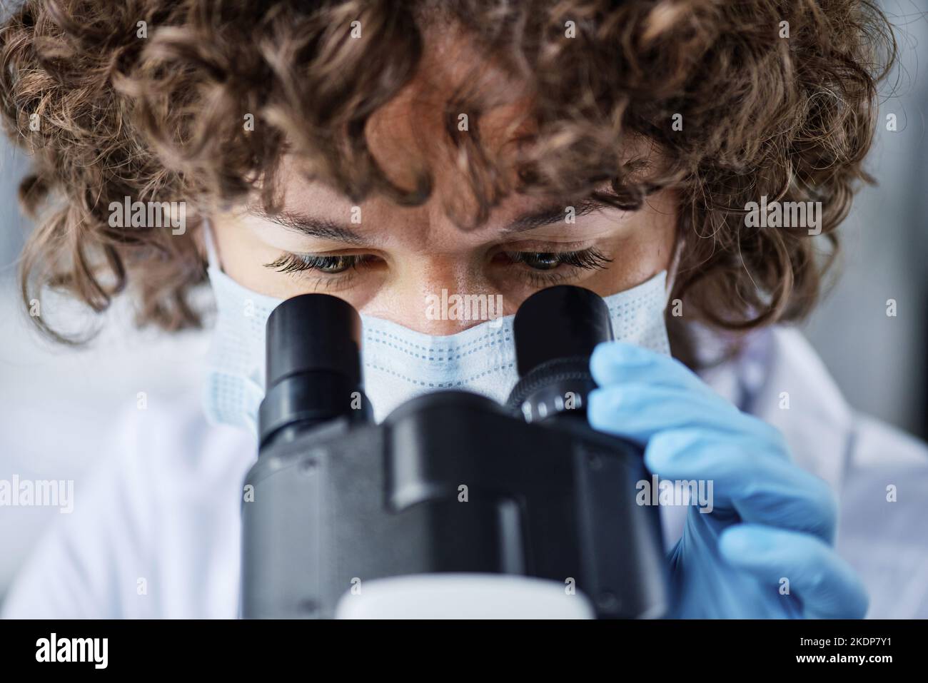 Nahaufnahme einer jungen Mikrobiologin in Schutzmaske, die im Mikroskop schaut, während sie während der Studie die Eigenschaften des neuen Virus untersucht Stockfoto