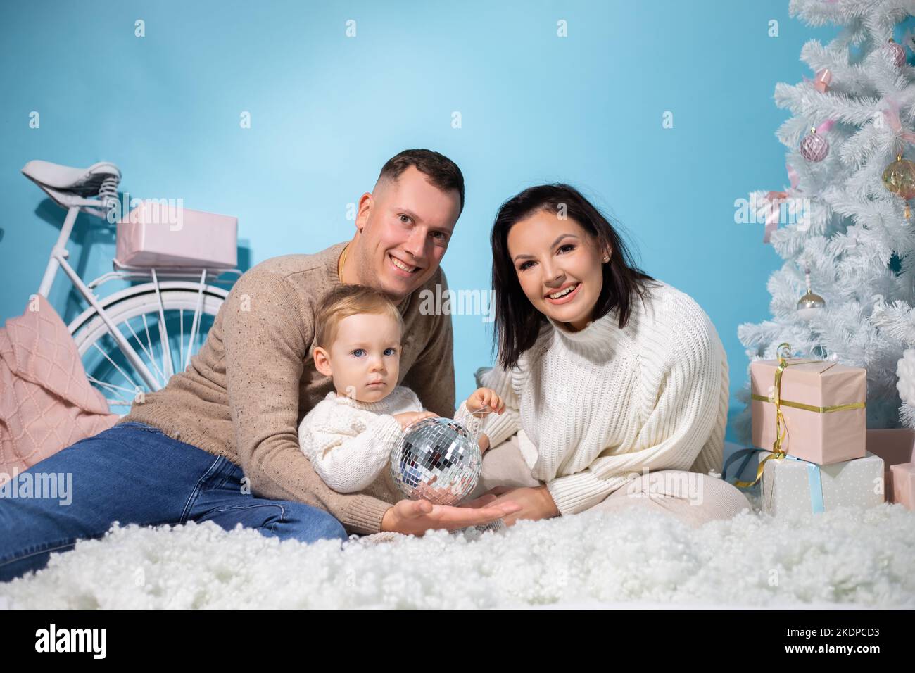 Familienportrait von drei Personen, die zusammen in einem weihnachtlichen Drehort mit weißen Dekorationen auf blauem Hintergrund sitzen. Glückliche Mutter, Vater und Stockfoto