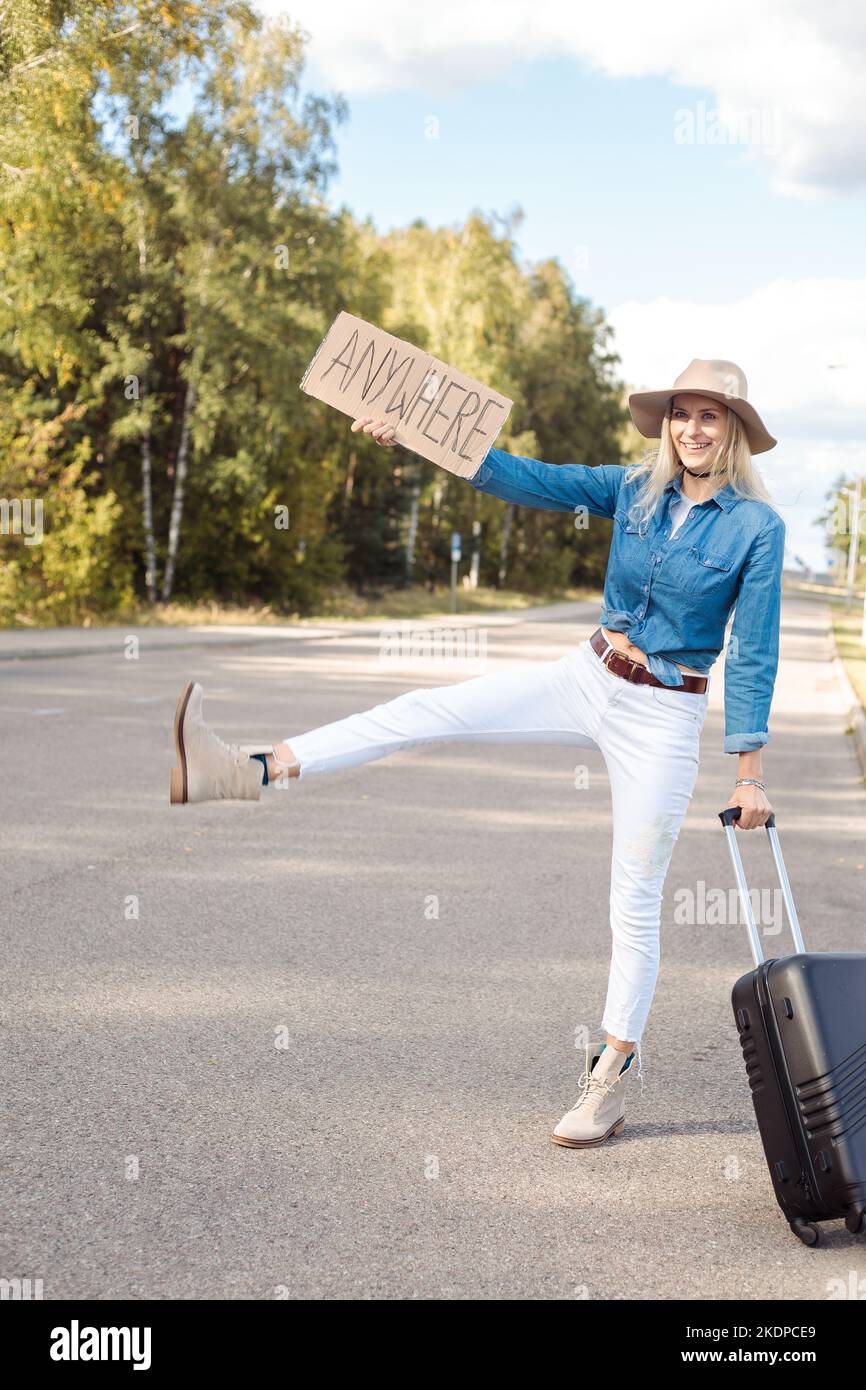 Vertikales Betteln lustig lächelnd blonde Frau Autostop Anhalter durch erhöhte Bein Zeichen, mit Teller überall Gepäck tragen Stockfoto