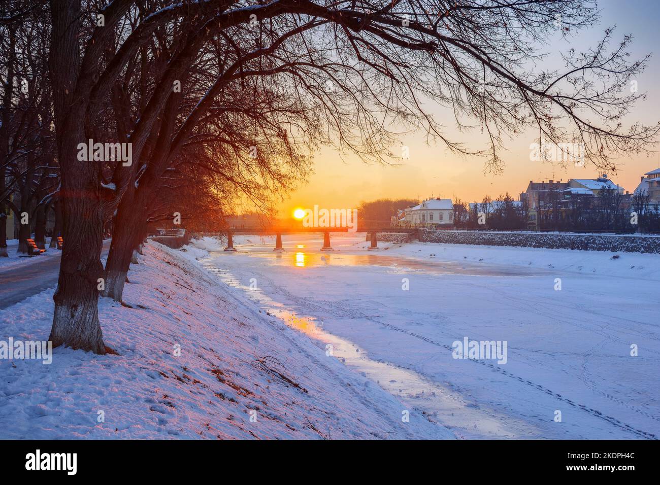 Wintersonnenaufgang in uzhgorod. Schöne Stadtlandschaft am frühen Morgen. Sonne über dem Horizont und Brücke über den gefrorenen Fluss. Schneebedeckte Böschung Stockfoto