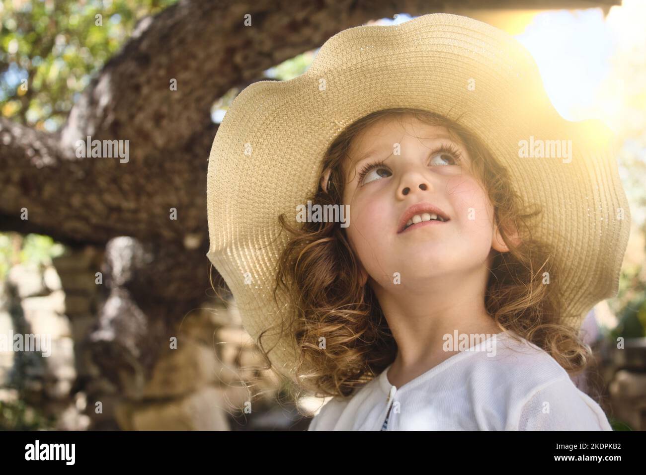 Nahaufnahme eines niedlichen kleinen Mädchens, das einen ländlichen Strohhut trägt und mit leuchtendem Sonnenlicht im Hintergrund aufschaut Stockfoto
