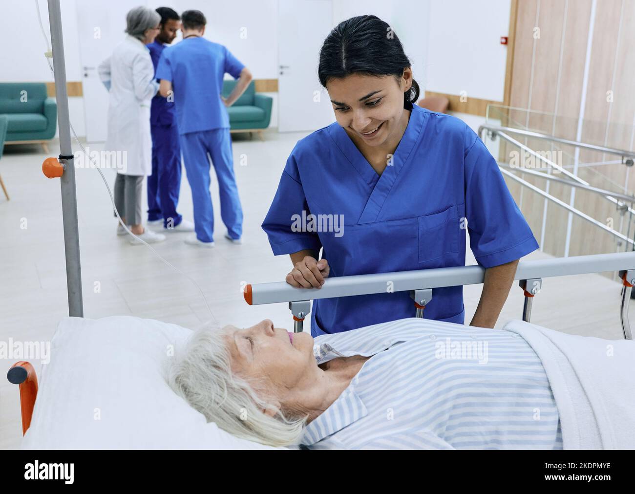 Positive Krankenschwester unterstützt kranke ältere Frauen, die auf der Liege im Krankenhauskorridor vor dem Hintergrund des ärzterates liegen. Patientenversorgung im Krankenhaus Stockfoto